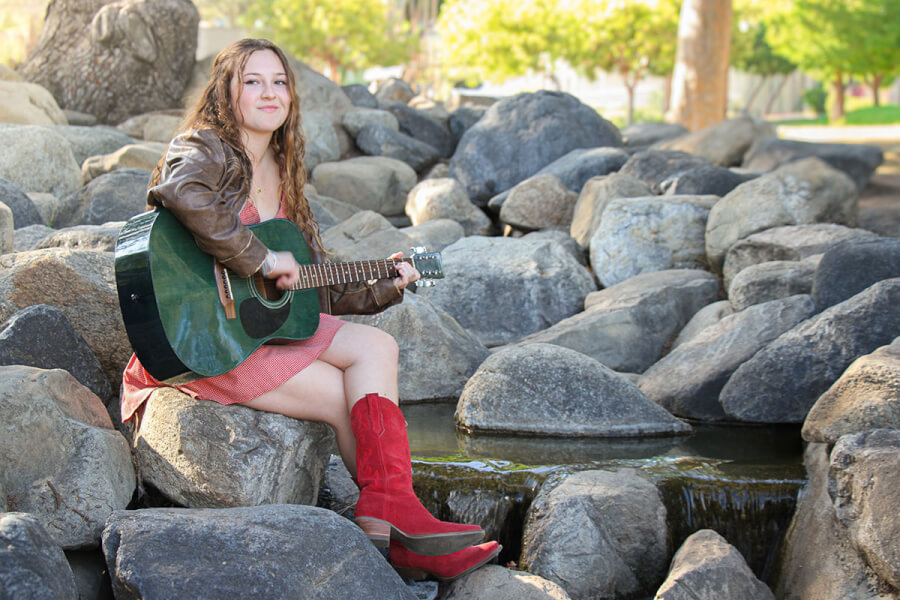 Young woman sits on large rocks by a small stream, playing a green acoustic guitar, wearing a brown leather jacket, pink dress, and red boots.
