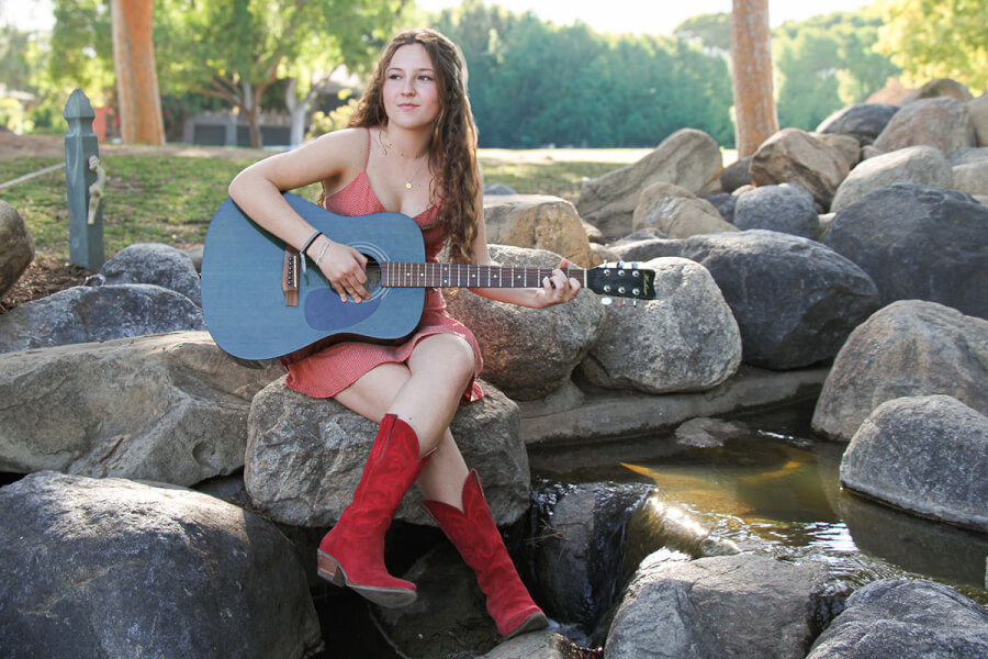 Young woman in a coral dress sits on rocks by a small stream, playing a blue acoustic guitar.