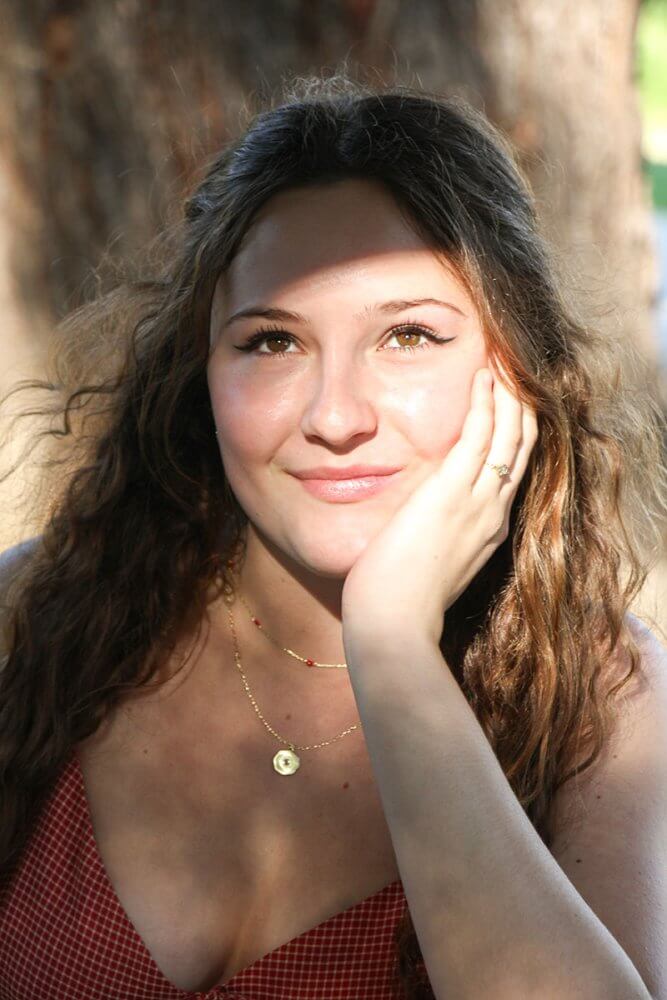 Portrait of a young woman with wavy hair, resting her head on her hand and smiling in sunlight outdoors, wearing a red checkered top.