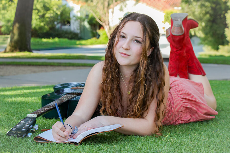 Young woman lying on grass in a park, writing in a notebook with a guitar beside her.