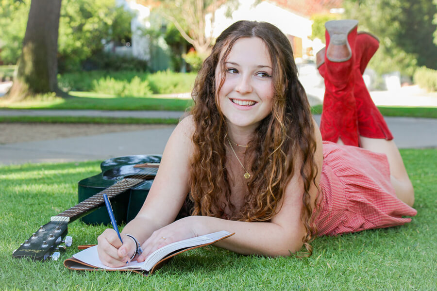 Young woman with long curly hair lying on grass in a park, writing in a notebook beside a guitar nearby.